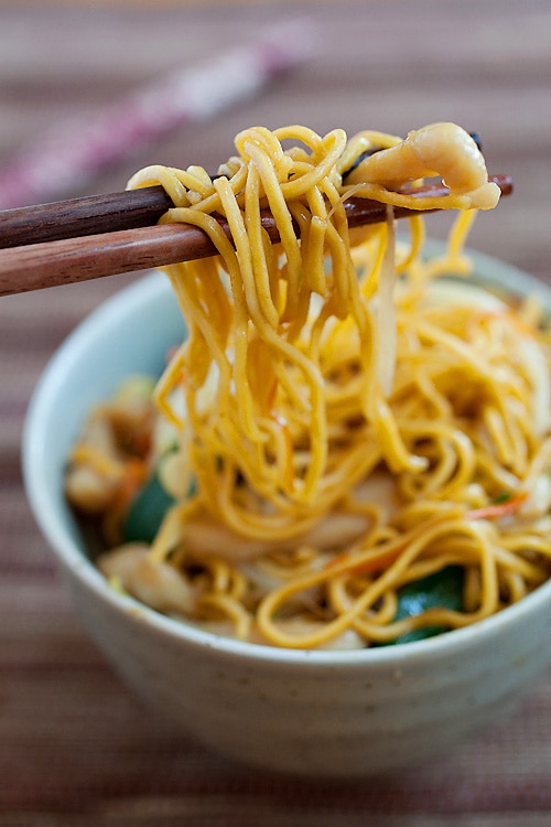 A pair of chopsticks holding up chicken chow mein with crispy noodles.
