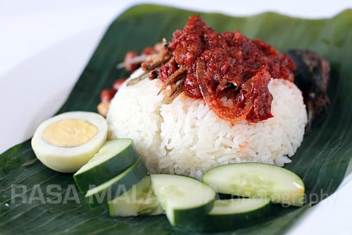 Close up of nasi lemak on a banana leaf with fried crispy anchovies, toasted peanuts, fried fish, sambal and cucumber.