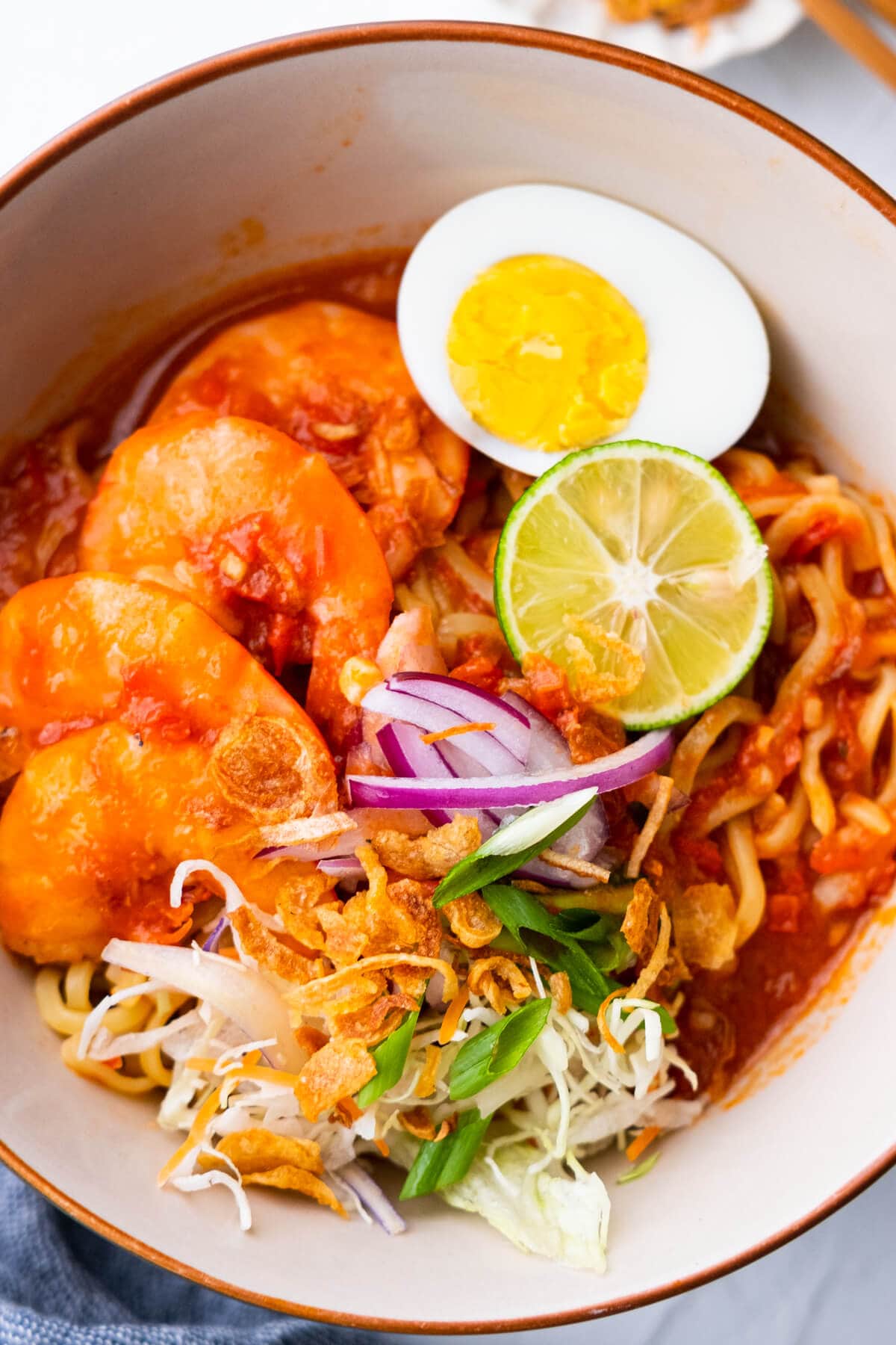 Malay prawn noodle served in a bowl with large prawns, boiled egg, lime, onion, spring onion and deep fried shallots on top. 