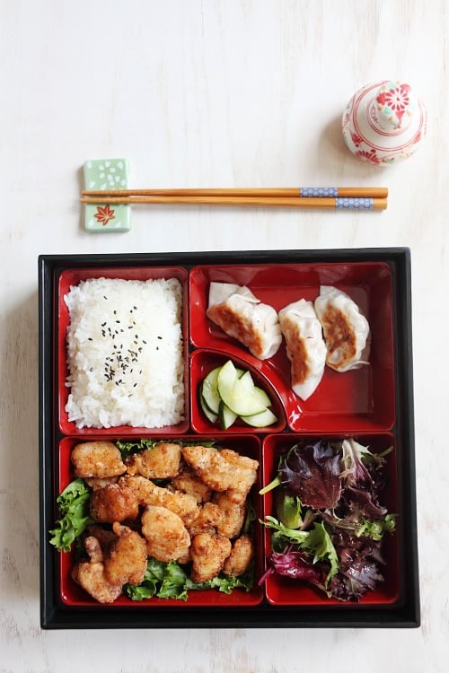 Tasty and quick Japanese fried chicken bento with steamed rice and sesame seeds, pickled cucumbers, salad and gyoza.