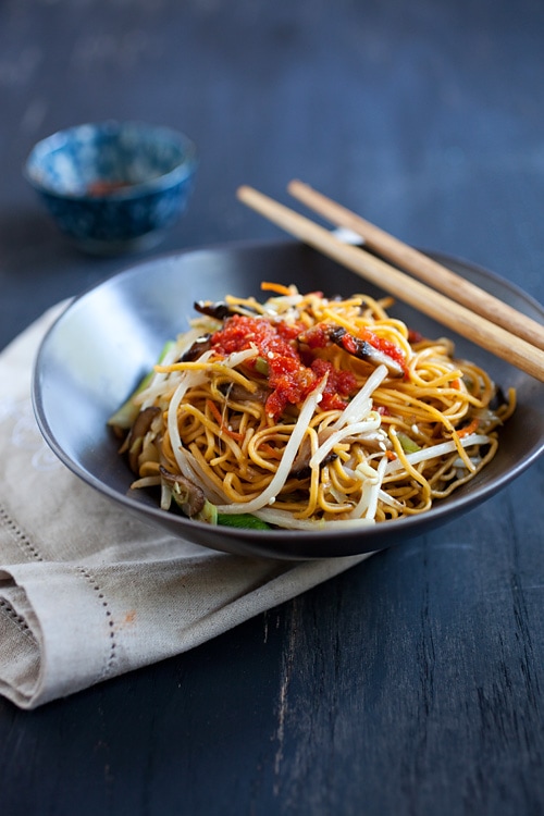 Bowl of Chinese vegetable stir fry noodles with red chili sauce and bean sprouts in bowl with chopsticks.