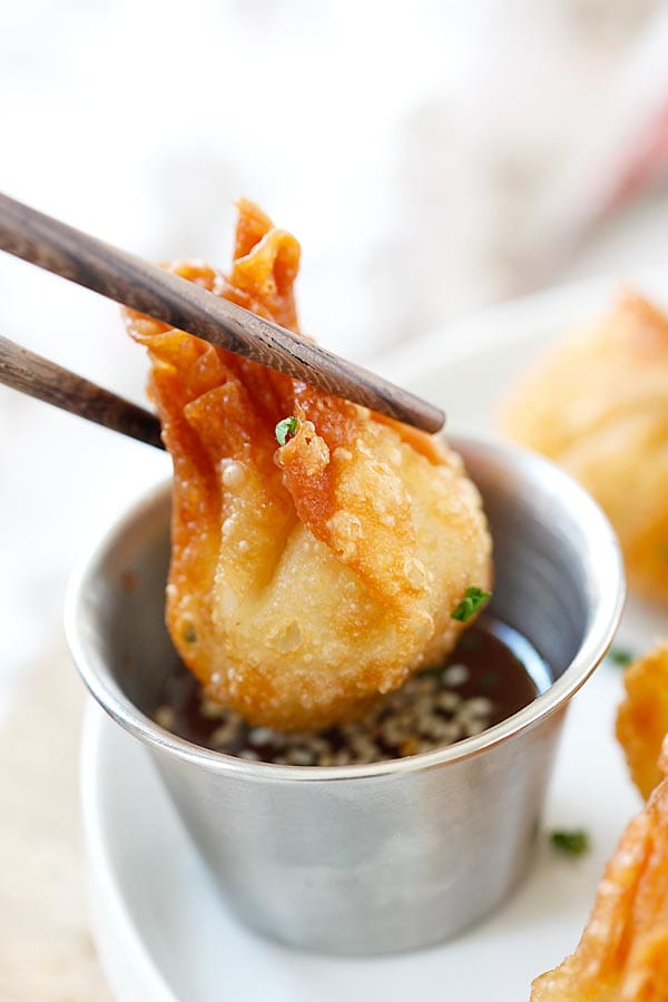 Small deep fried crispy wonton picked up by a pair of chopsticks dipping onto Asian wonton sauce.