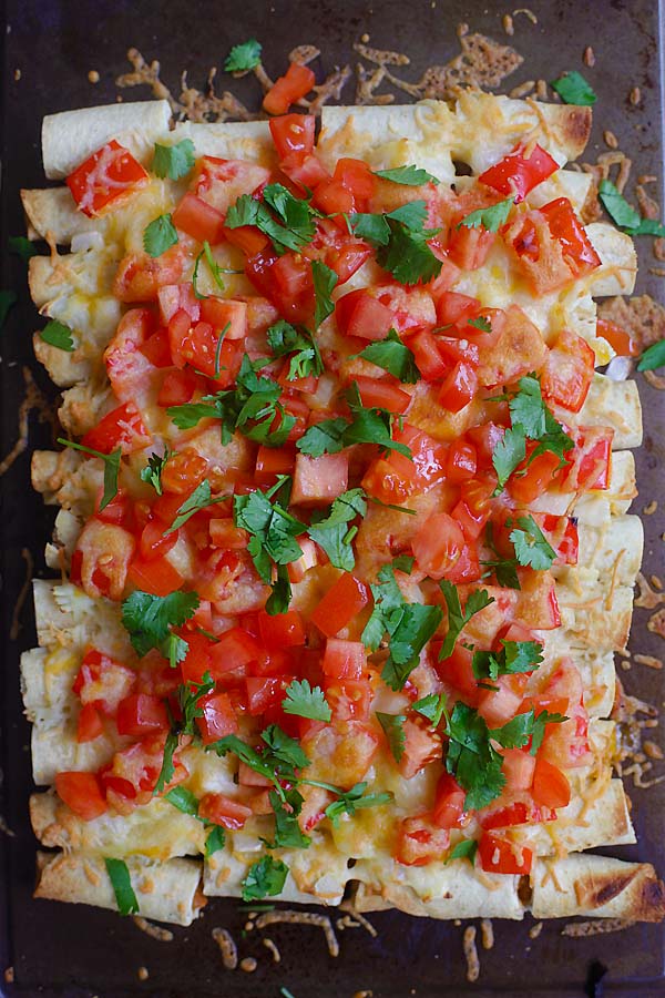 Easy and quick baked Mexican cheesy taquitos topped with chopped tomatoes, diced onions, bell peppers and shredded cheese on a baking tray.