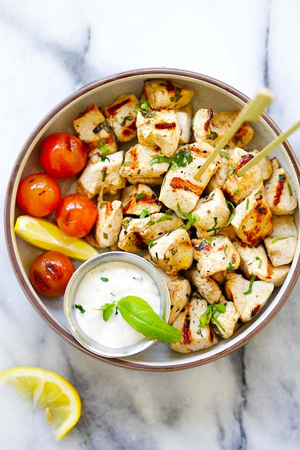 Char grilled Mediterranean chicken bites with Mediterranean marinade in a serving dish, with a side of grilled tomatoes and ranch dipping sauce.