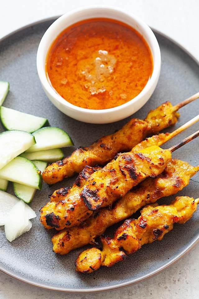 Five skewers of chicken satay on a plate with cucumber slices and satay sauce.