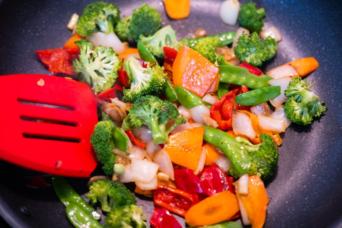 Stir-fry red bell peppers, carrot slices, onion, and broccoli in a frying-pan. 