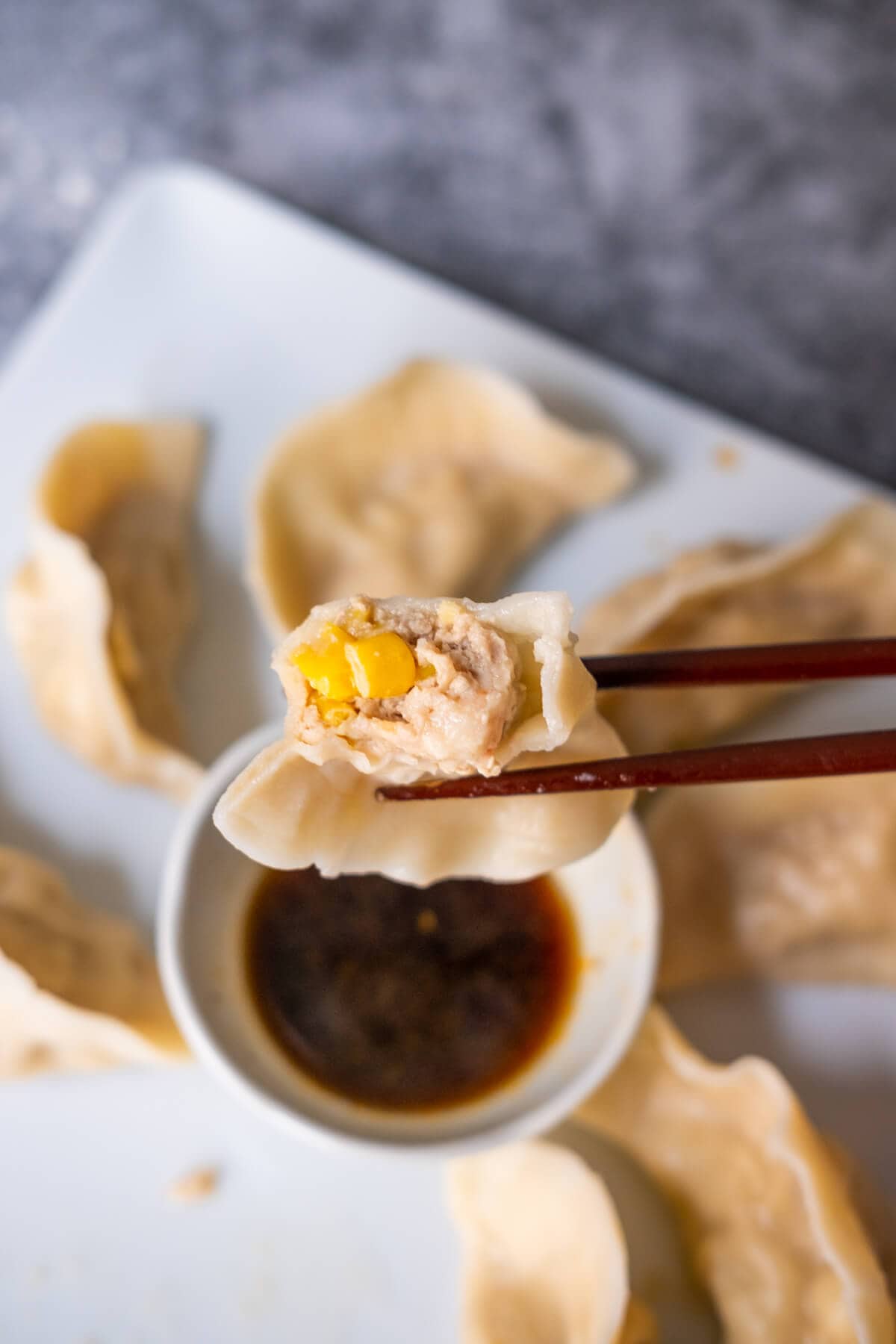 A half cut dumpling revealing its pork, shrimp, and sweet corn filling being held by a chopstick over a bowl of vinegar sitting in the center of a plate of dumplings. 