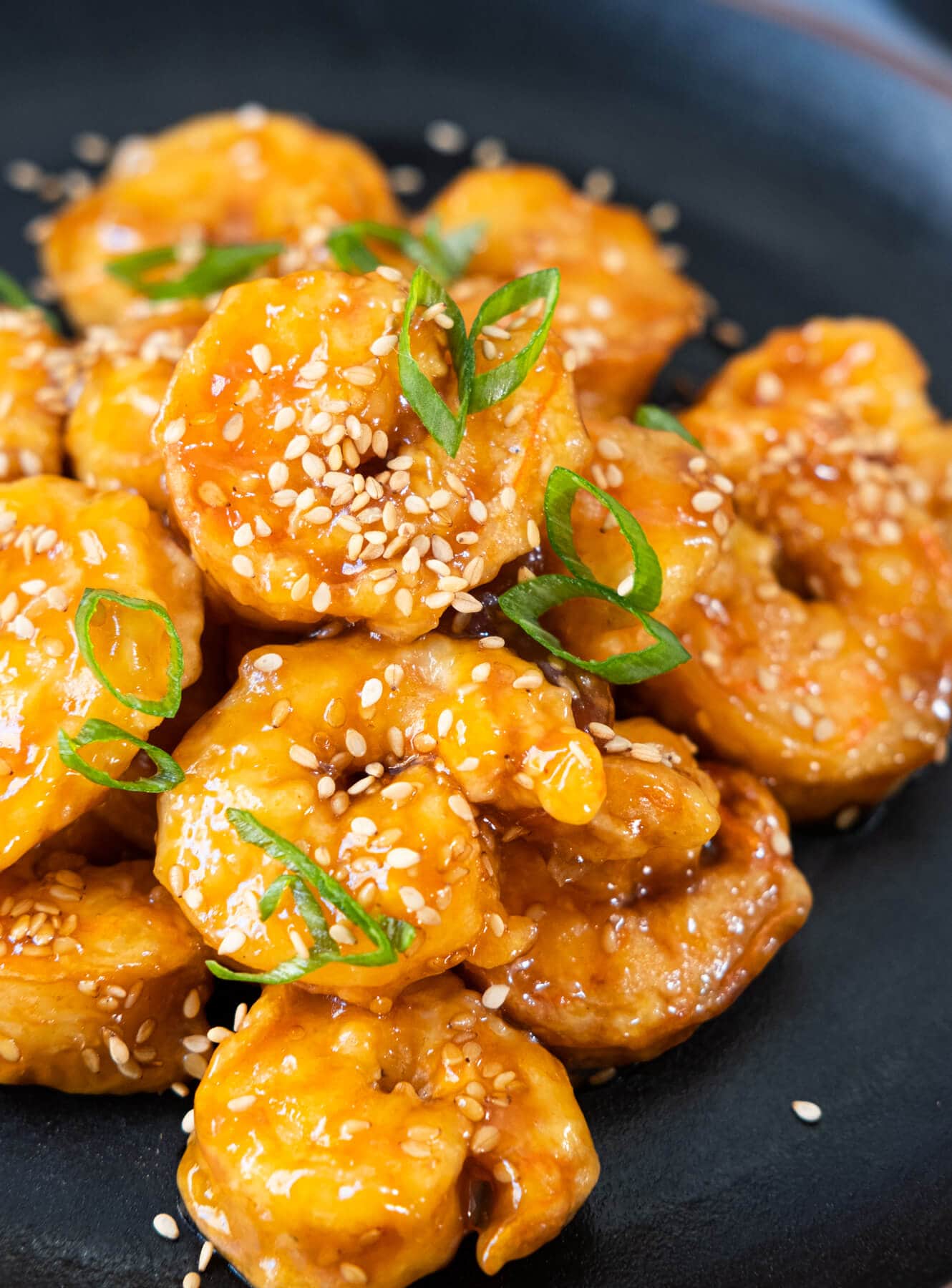 Shrimps coated with sweet orange sauce and  sesame seeds served on a black plate. 