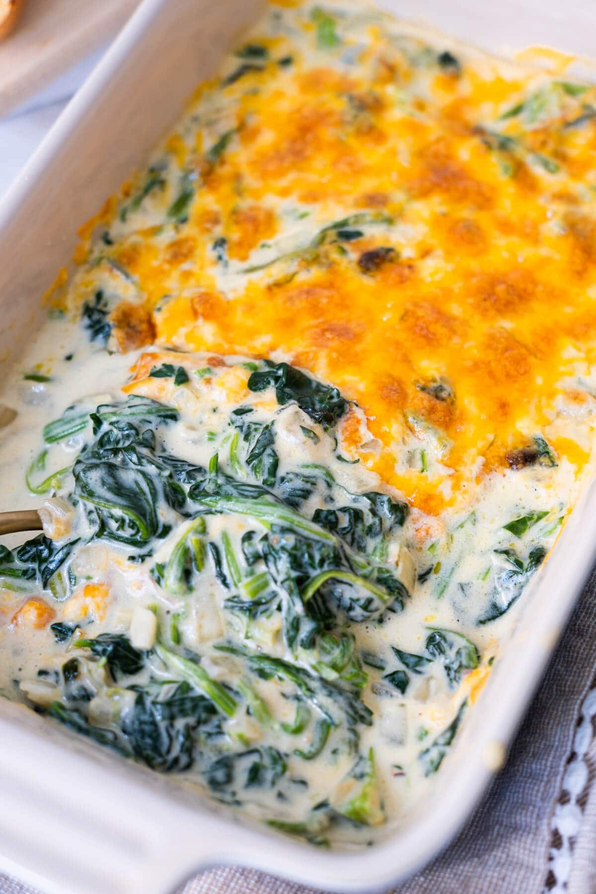 Creamed spinach is half topped with baked cheese in a baking dish with a kitchen towel under it.