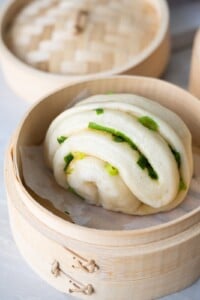 One scallion bun in a parchment lined bamboo steamer with the lid placed placed next to it.