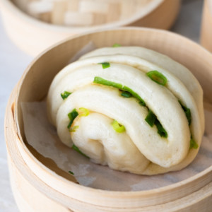 One scallion bun in a parchment lined bamboo steamer with the lid placed placed next to it.