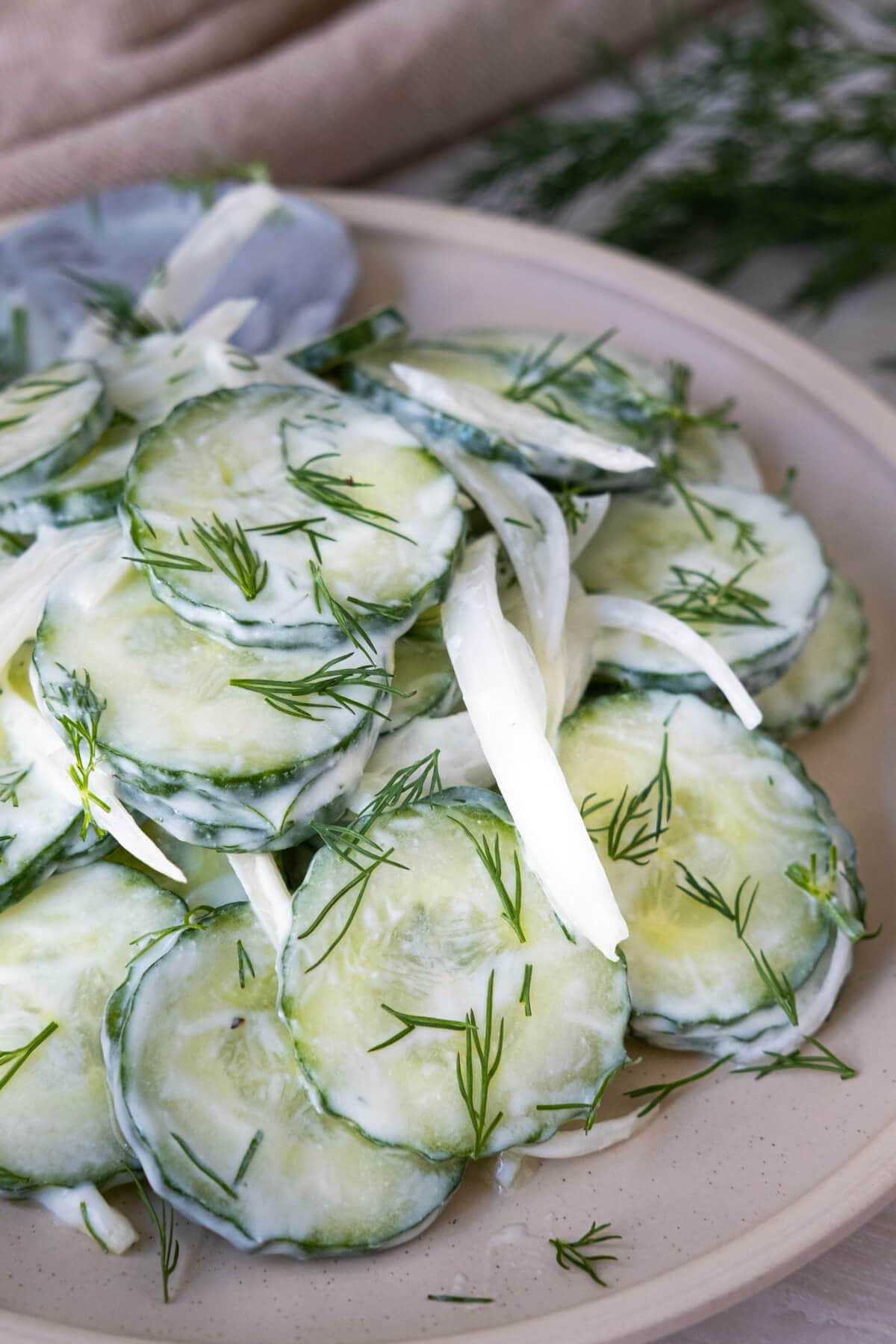 Slices of cucumbers dressed with Greek yogurt balsamic dressing served white onion slices and fresh chopped dill on top served in a pink plate.