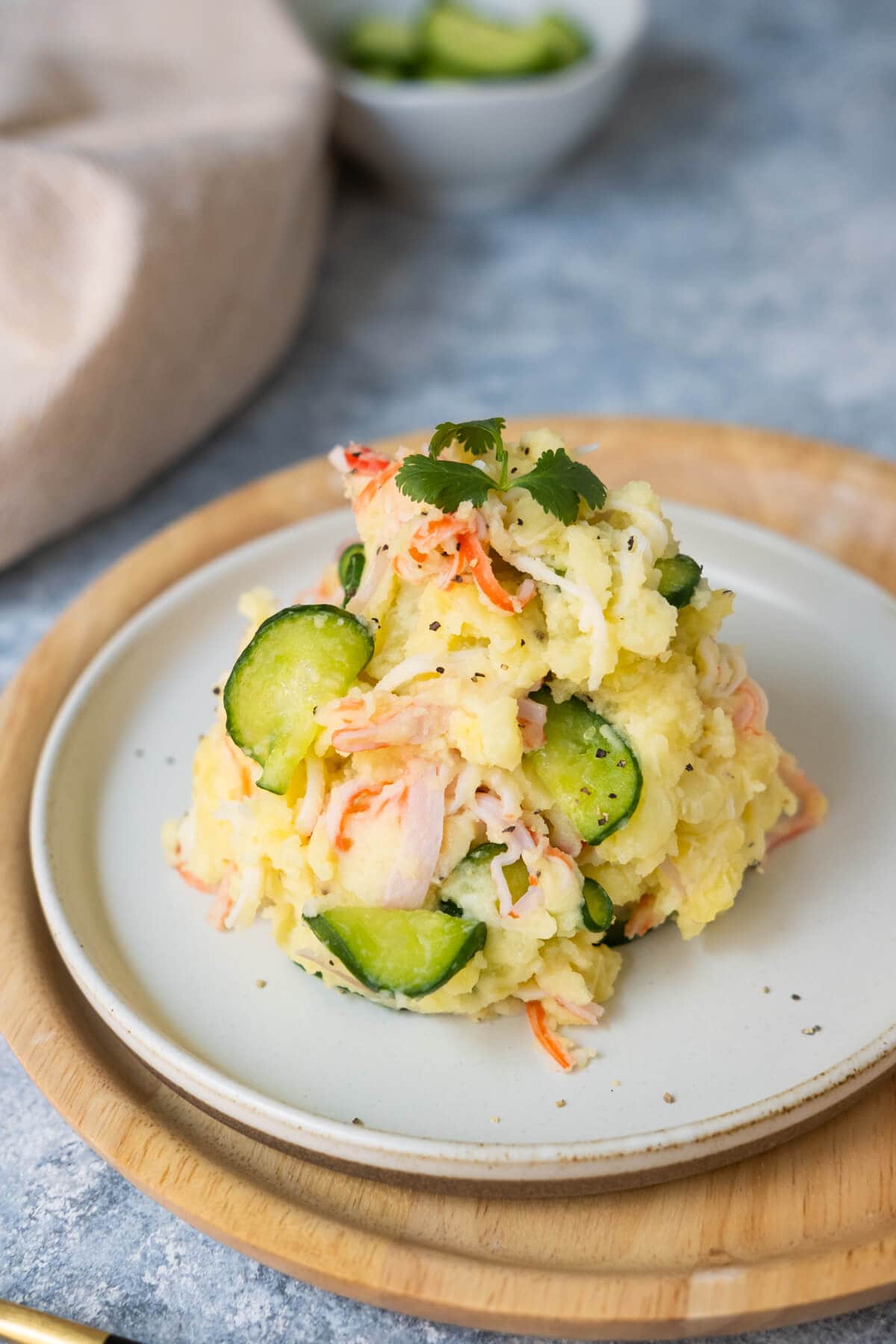 Potato salad containing Japanese cucumber slices and sliced imitation crab meat on a plate with kitchen towel and a small side of cucumber slices placed behind. 