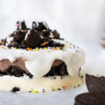 Ice cream cake topped with Oreo crumbs and sprinkles with whipped cream drizzle down the side.