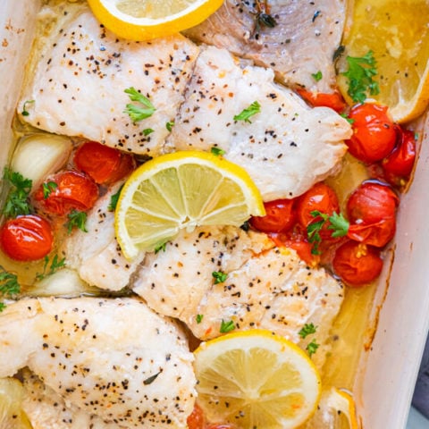 Baked catfish with cherry tomatoes, lemon slices and parsley in a baking dish.