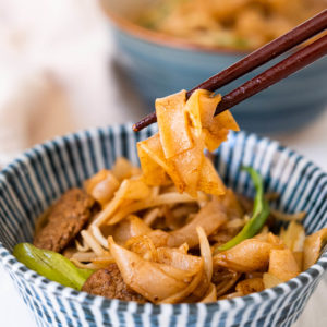 Beef chow fun noodles served in a blue bowl and picked up with a pair of chopsticks.