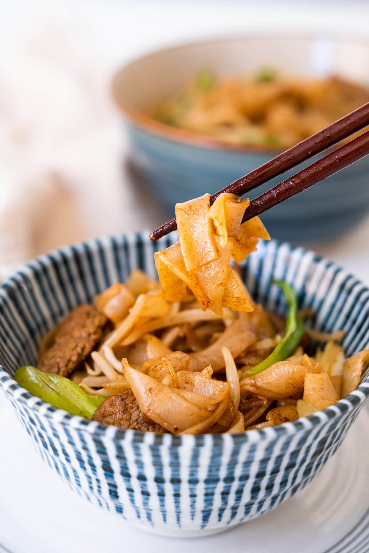 Beef chow fun noodles served in a blue bowl and picked up with a pair of chopsticks.