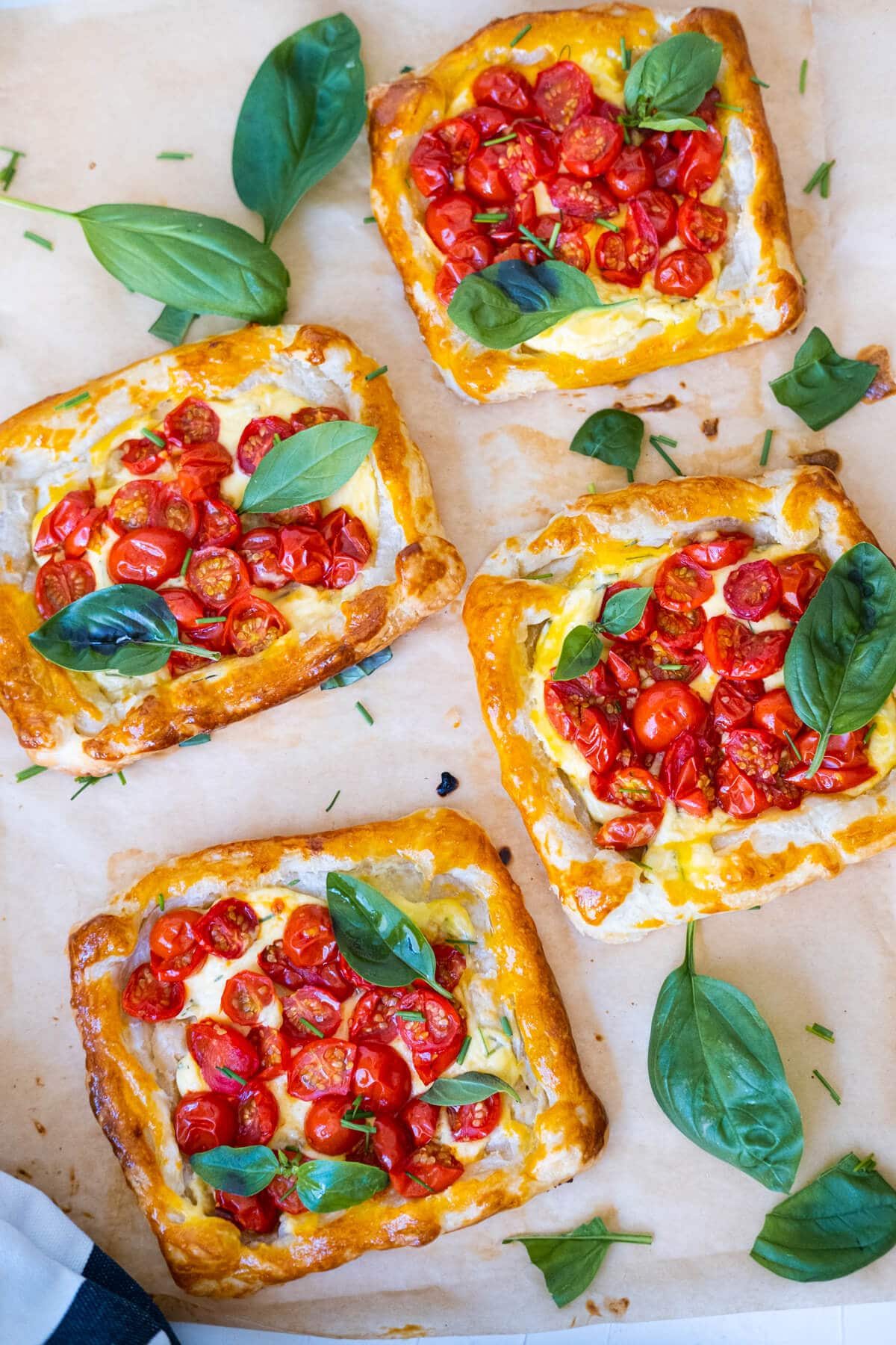 Four small rectangle cherry tomato tarts on a parchment paper and topped with fresh basil leaves. 