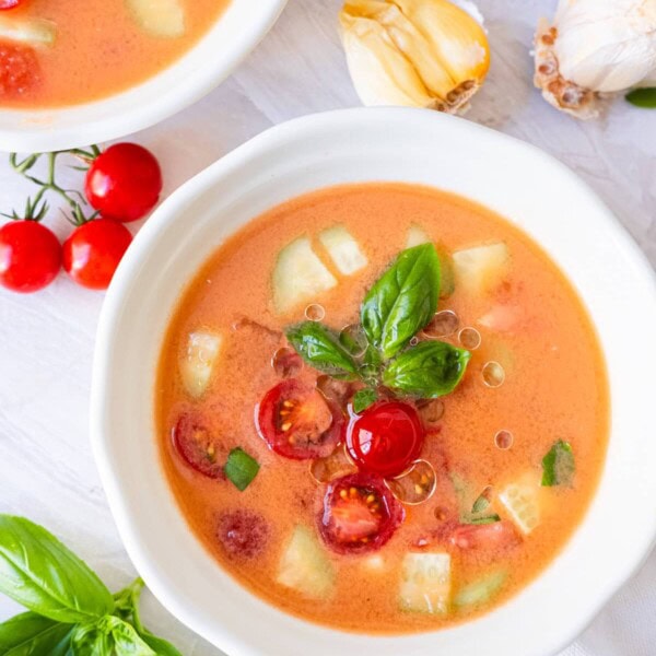 Easy gazpacho topped with cherry tomato and cucumber chunks in a small bowl with garlic and basil leaves on the side.