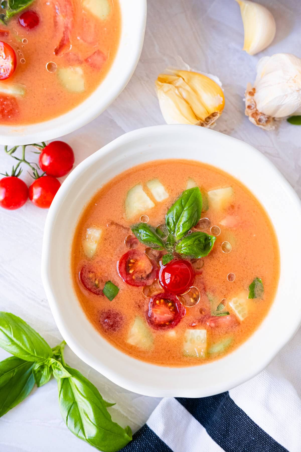 Easy gazpacho topped with cherry tomato and cucumber chunks in a small bowl with garlic and basil leaves on the side.
