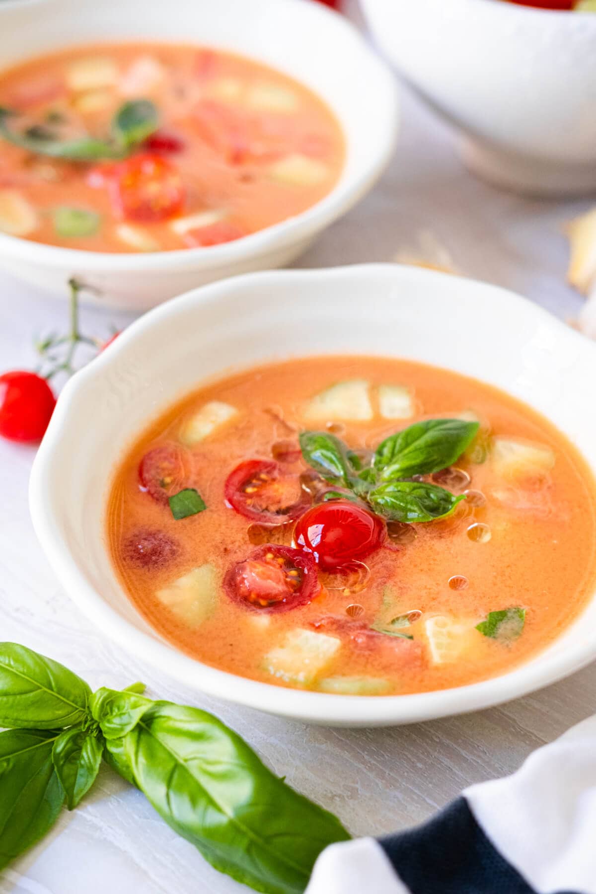 Orange-red easy gazpacho served in a white small bowl with tomatoes, cucumber chunks and basil leaves in top. 