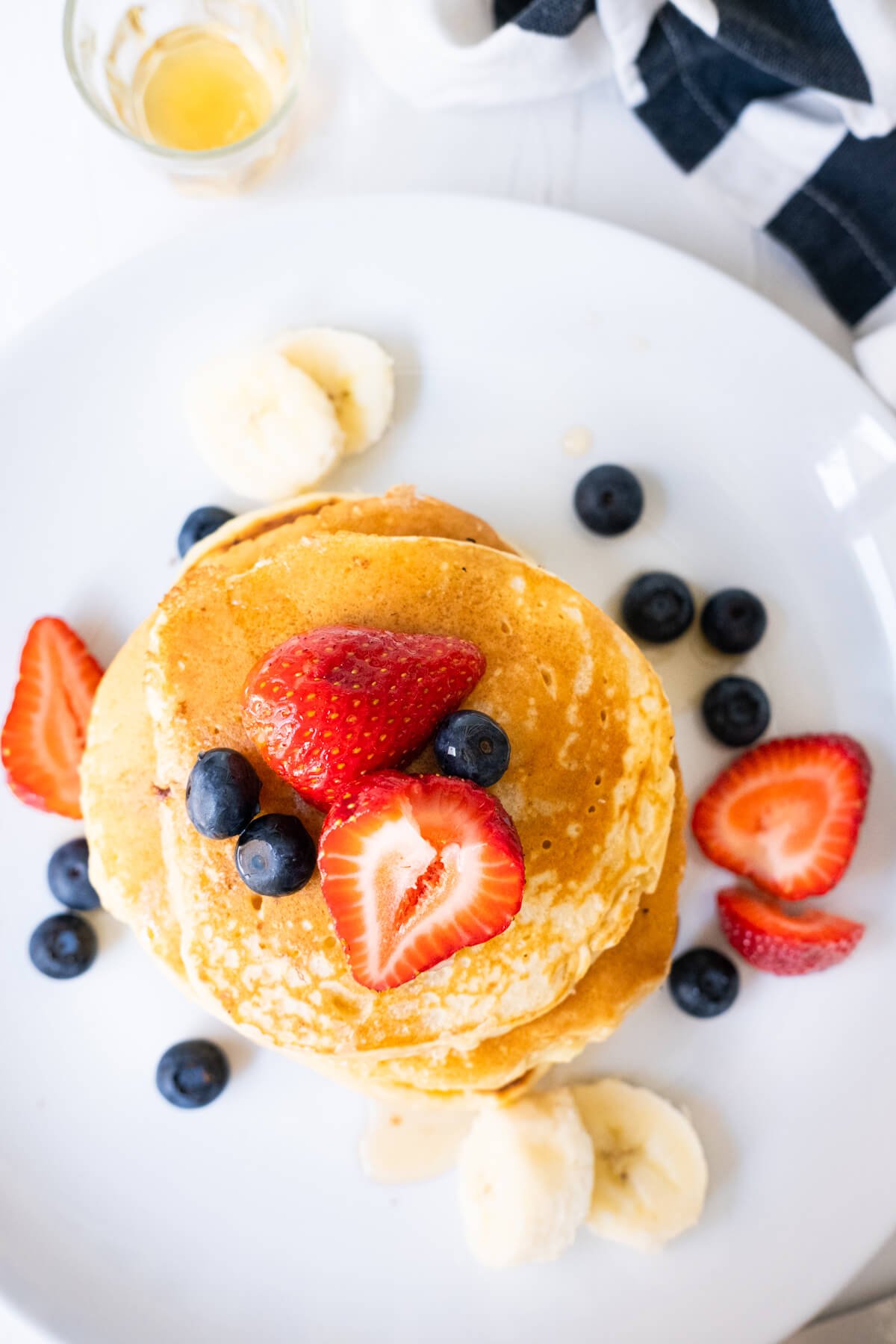 Fluffy pancakes in a white plate with blueberries, strawberries and banana slices on top and are served with maple syrup aside. 