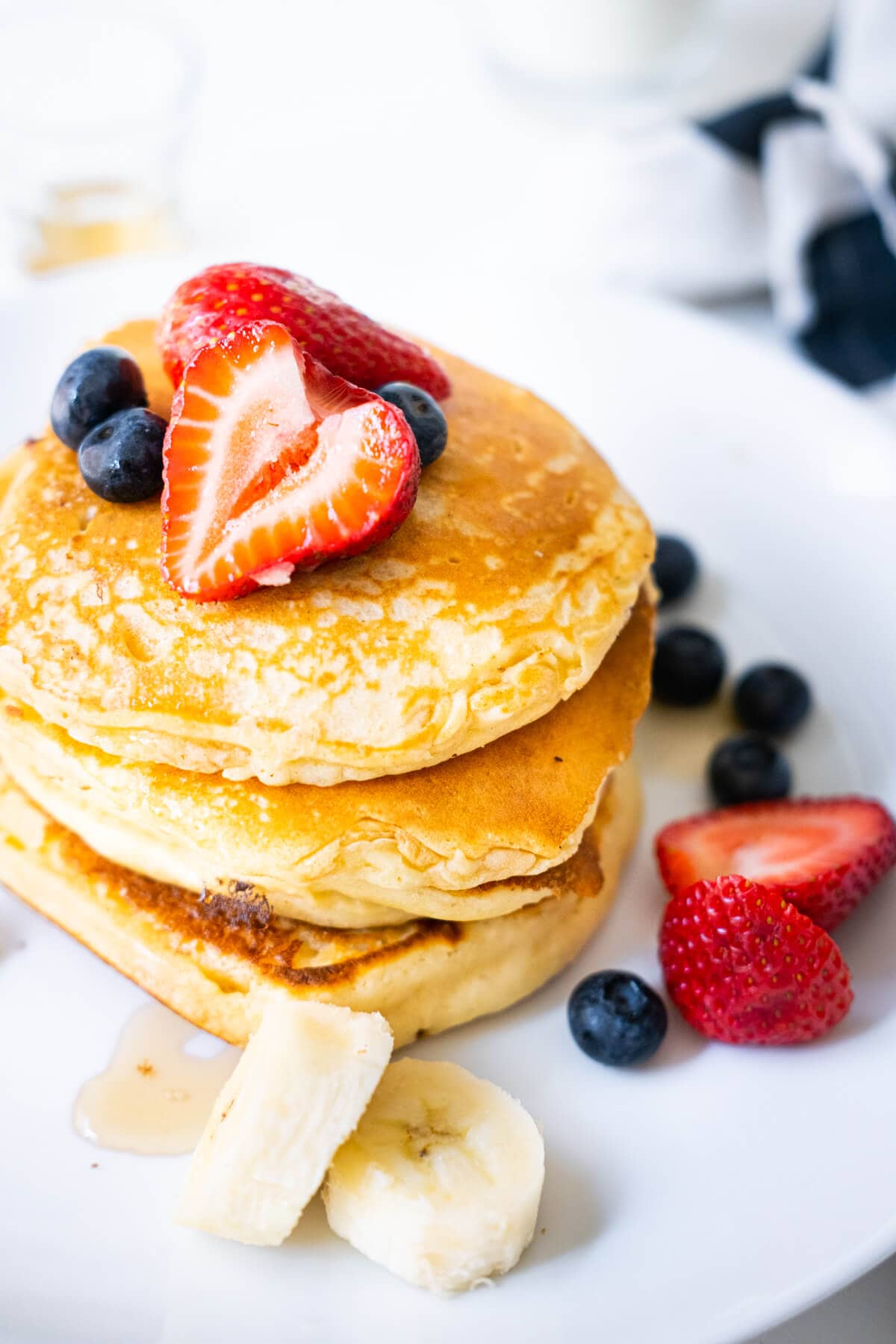 A stack of delicious pancakes served with maple syrup, strawberries, blueberries and slices of banana in a plate with kitchen towel on the side. 