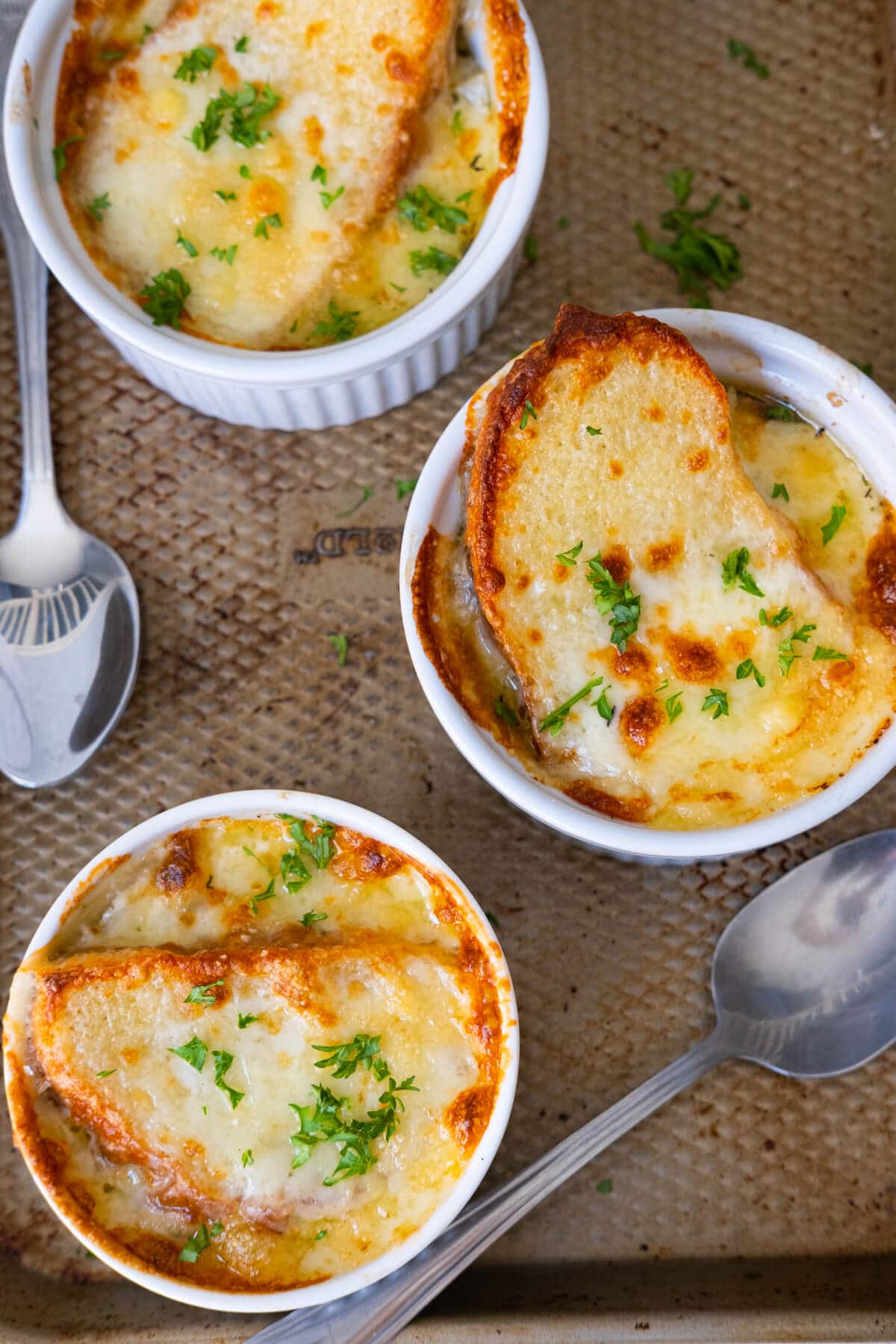 Three easy French onion soup served in ramekins and topped with bread and melted cheese, and placed on a baking pan with two spoons aside. 
