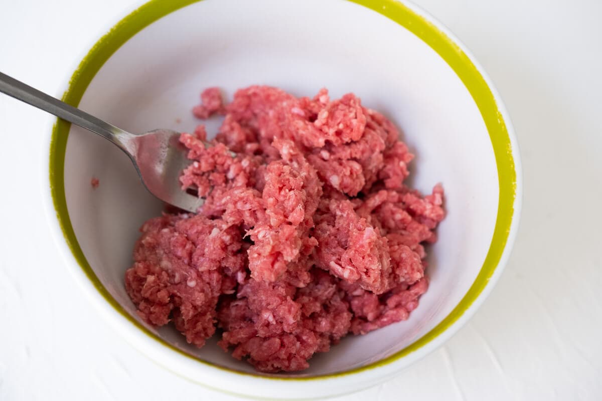 Fork braking up the ground beef into small pieces in a bowl. 