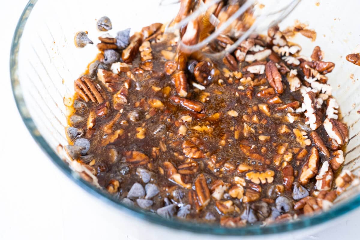 Chopped pecans and semisweet chocolate chips mixed together with the combination of eggs, maple syrup, brown sugar, vanilla extract, melted butter, and sea salt in a bowl. 