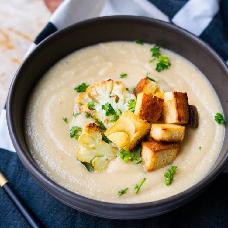 Creamy cauliflower soup in a soup bowl with 2 small roasted cauliflower florets, croutons and chopped parsley on top.