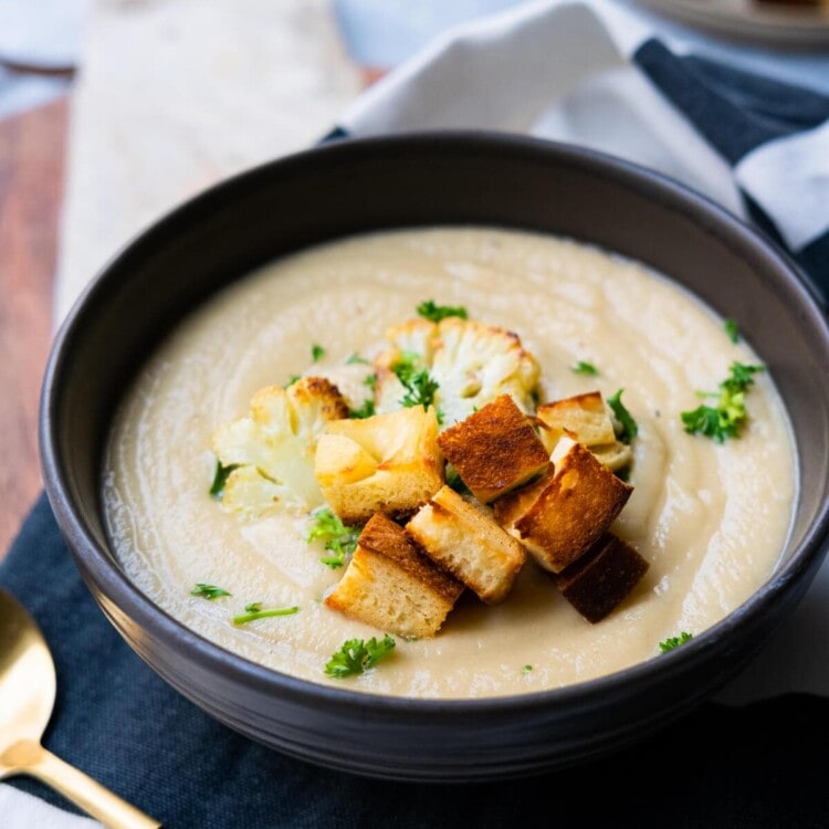 Creamy cauliflower soup in a bowl and placed on a wooden plate board with extra croutons on the side.