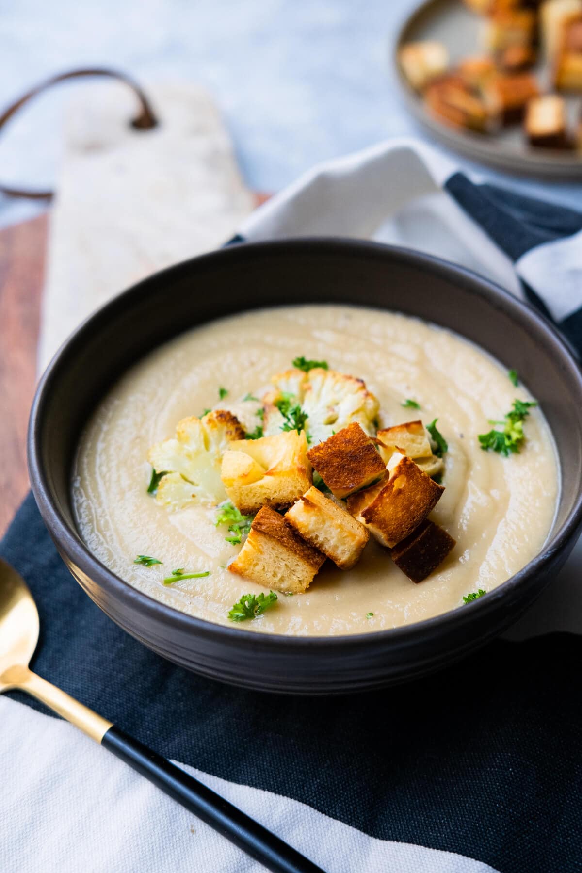 Soup in a bowl filled with cauliflower and croutons on a wooden plate board with extra croutons on the side. 
