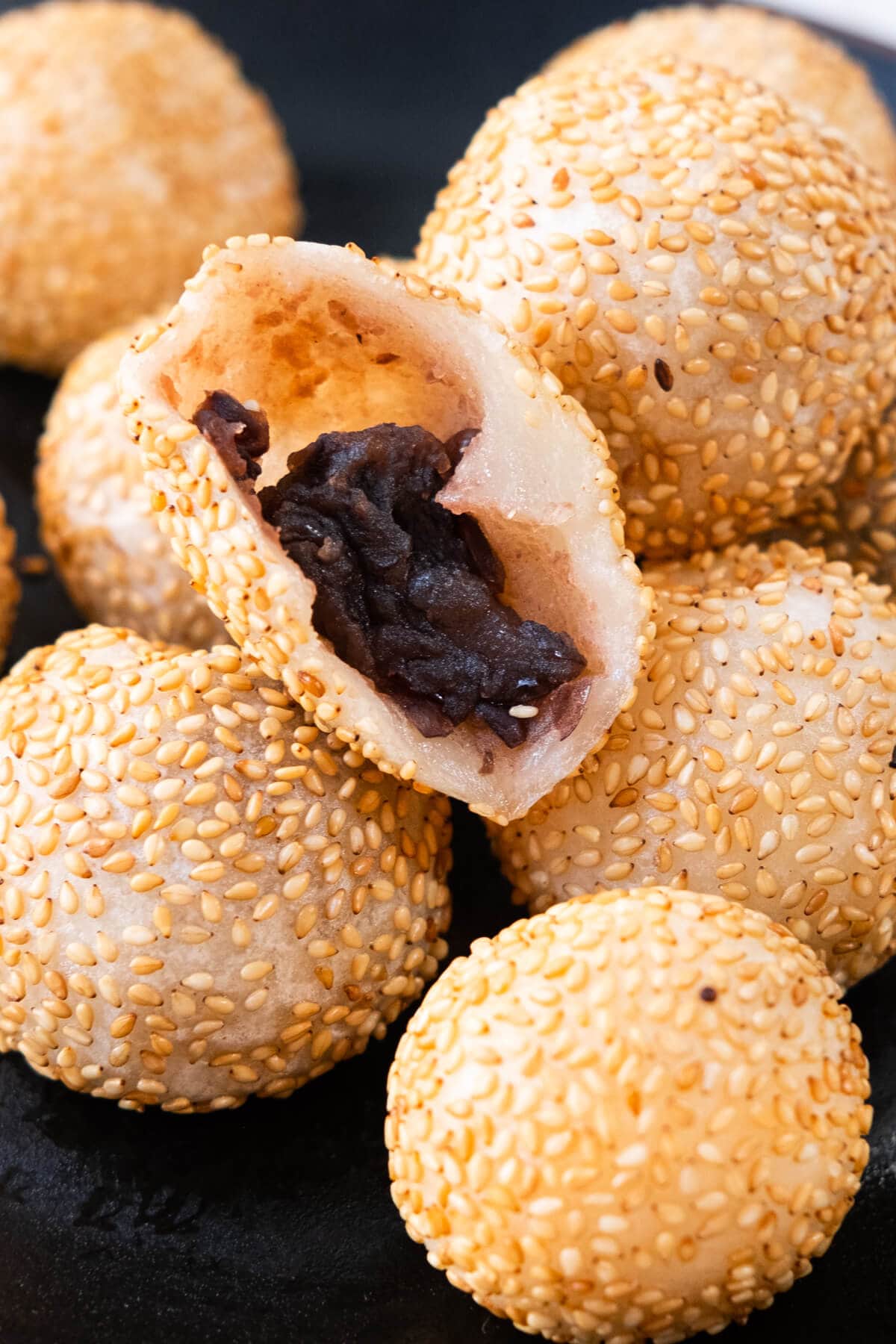 A half opened sesame coated ball with red bean paste filling inside surrounded by multiple sesame balls. 