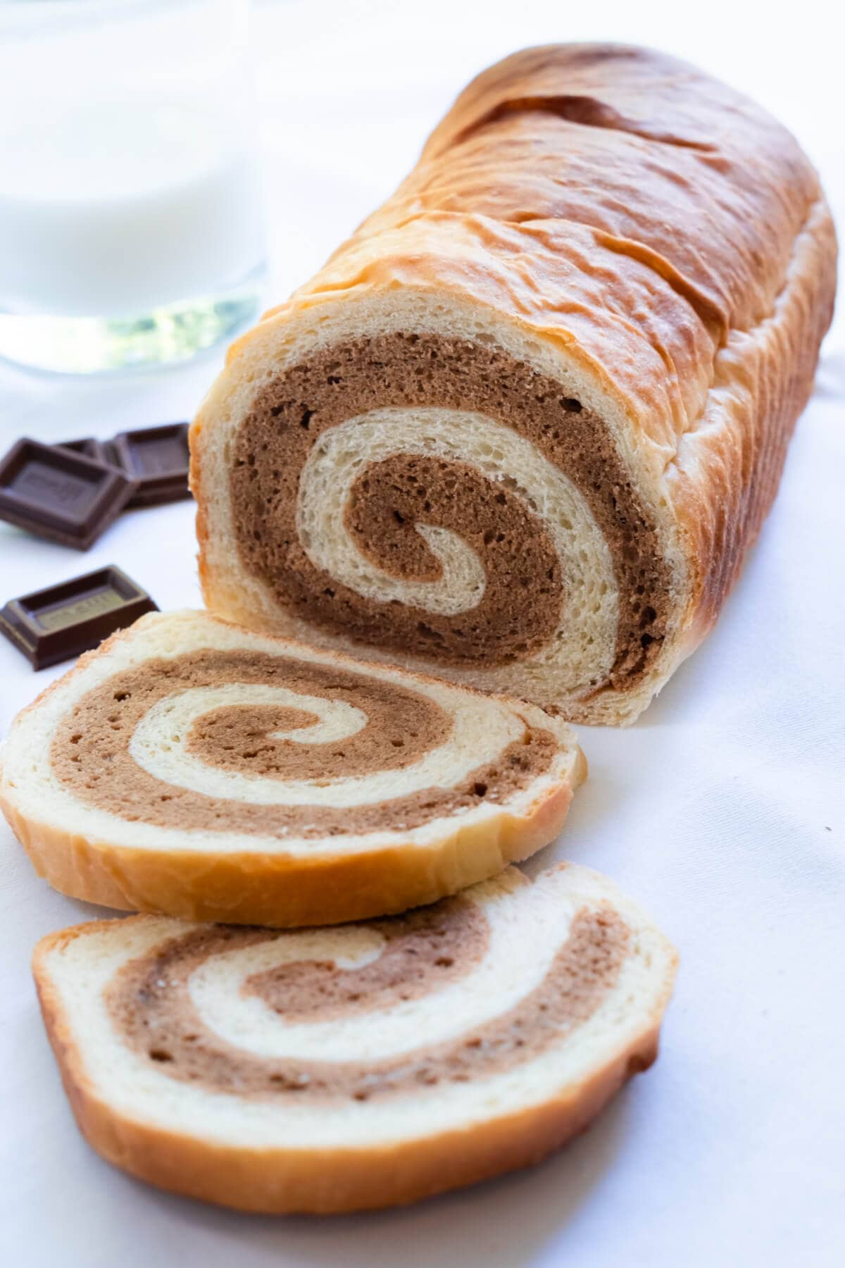 A loaf of cake bread in chocolate flavor with a cup of milk and few pieces of chocolate on the side. 