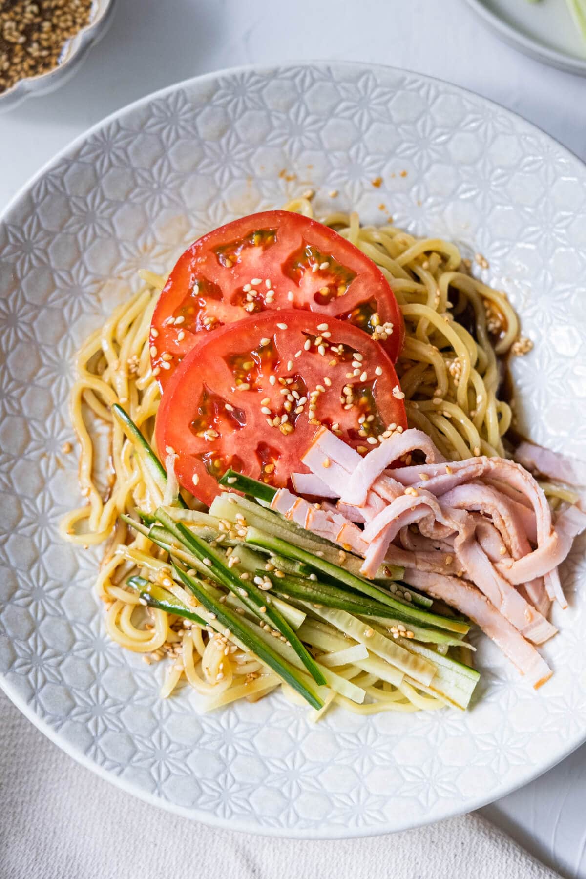Refreshing cold sesame noodle served in a white shallow plate with tomato, cucumber and ham. 