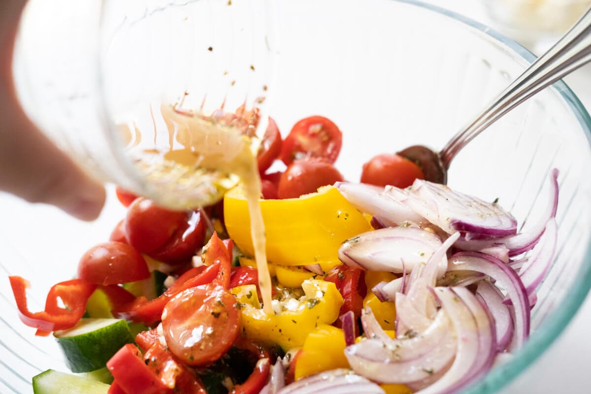 Vinaigrette poured over cucumber, red bell pepper, yellow bell pepper, cherry tomatoes, and red onions in a large mixing bowl.  