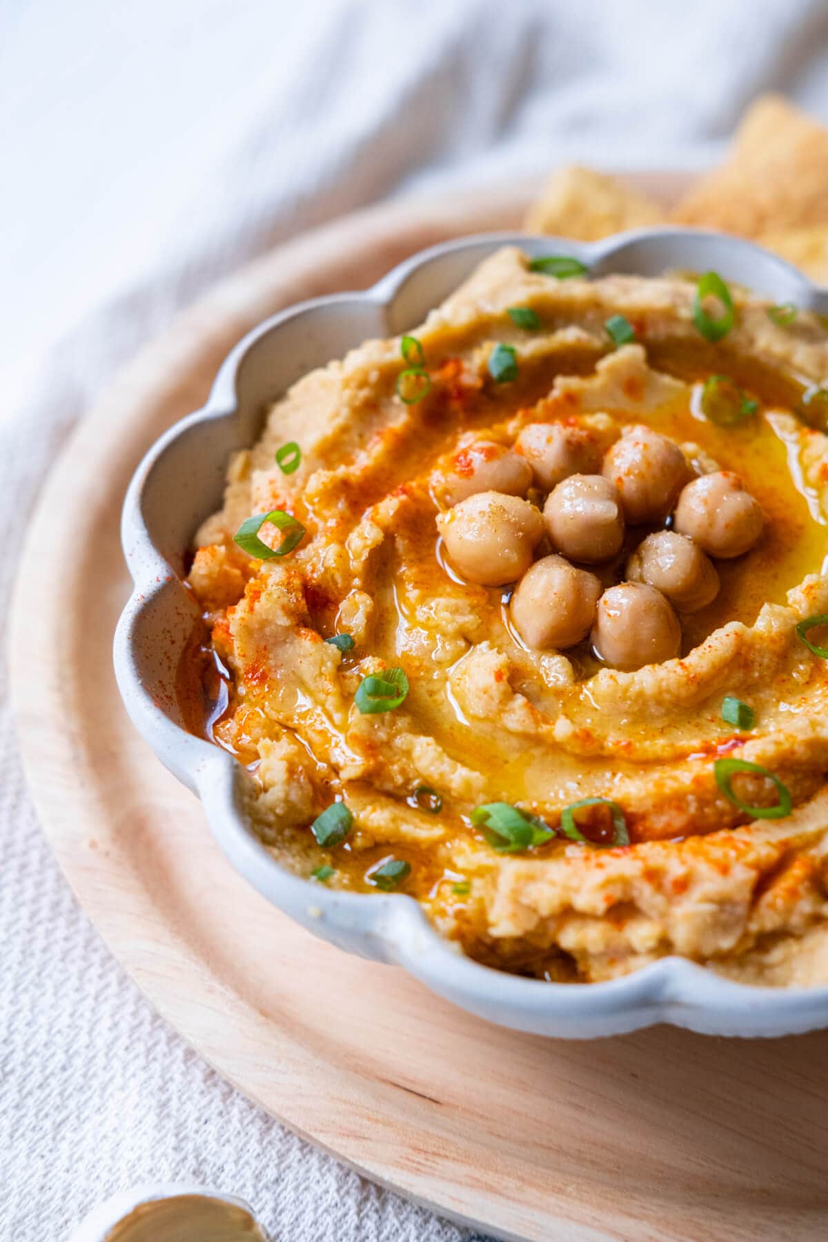 Close up shot of smooth hummus sprinkled with paprika and freshly chopped green onion with chickpeas on top drizzled with olive oil served in a bowl. 