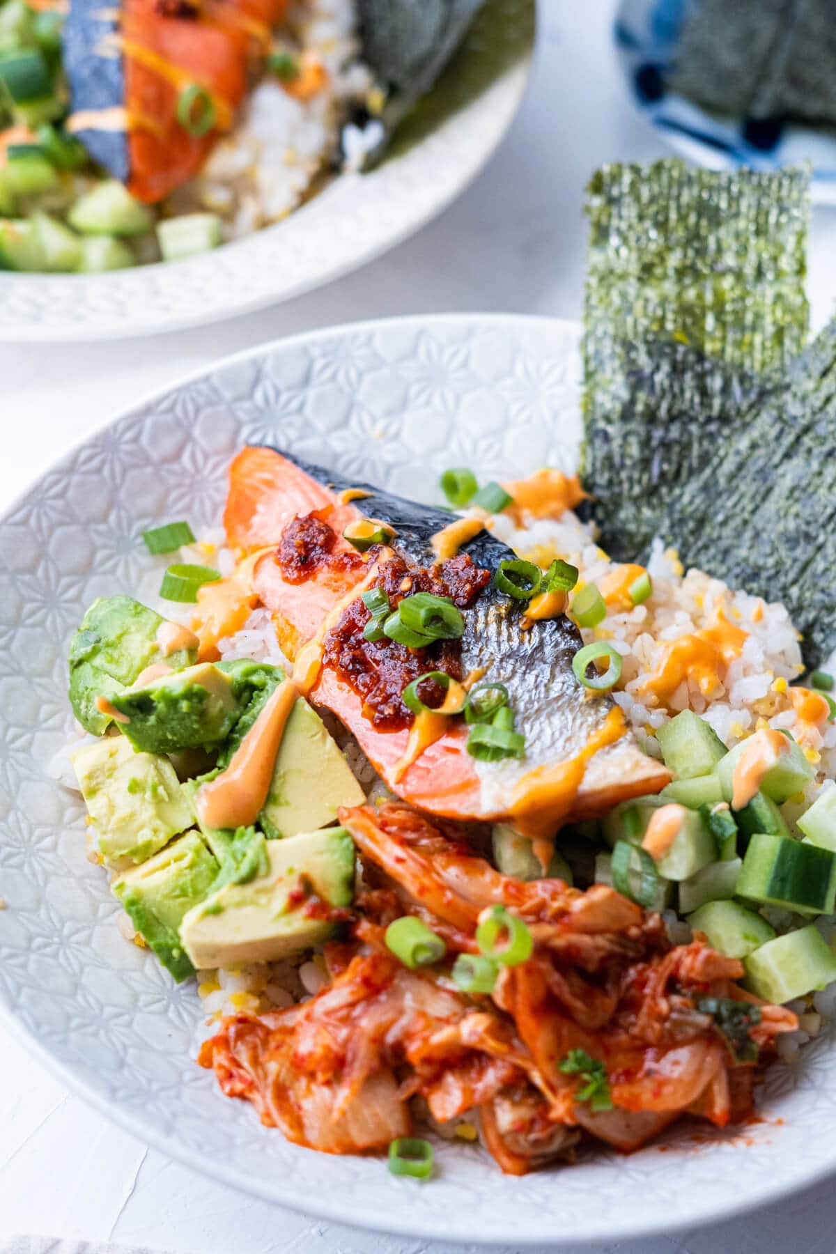 Close up of a bowl filled with cooked salmon over brown rice alongside avocado, cucumber, kimchi and nori. 