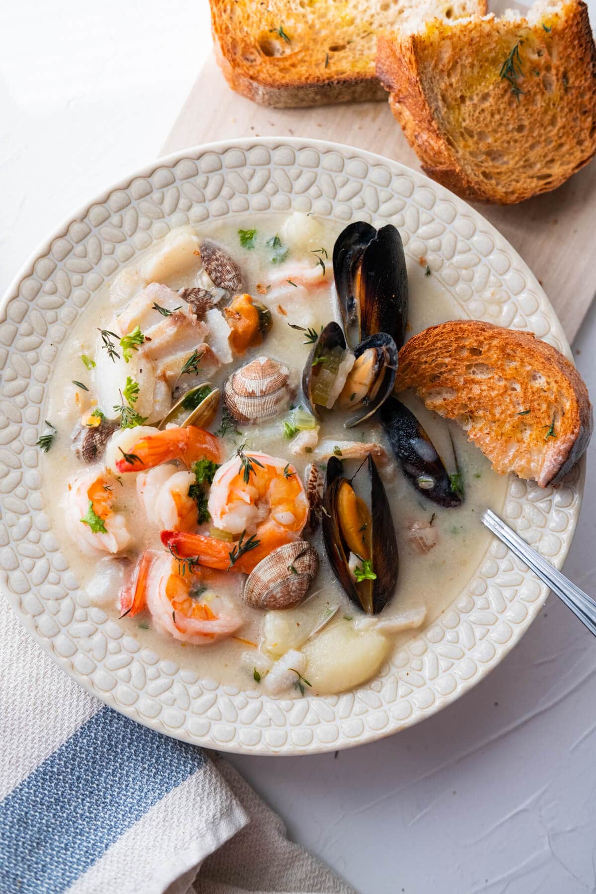 A plate of a creamy chowder with tons of seafood including clams, mussels, prawns, and fish, and alongside toasted sourdough slices. 