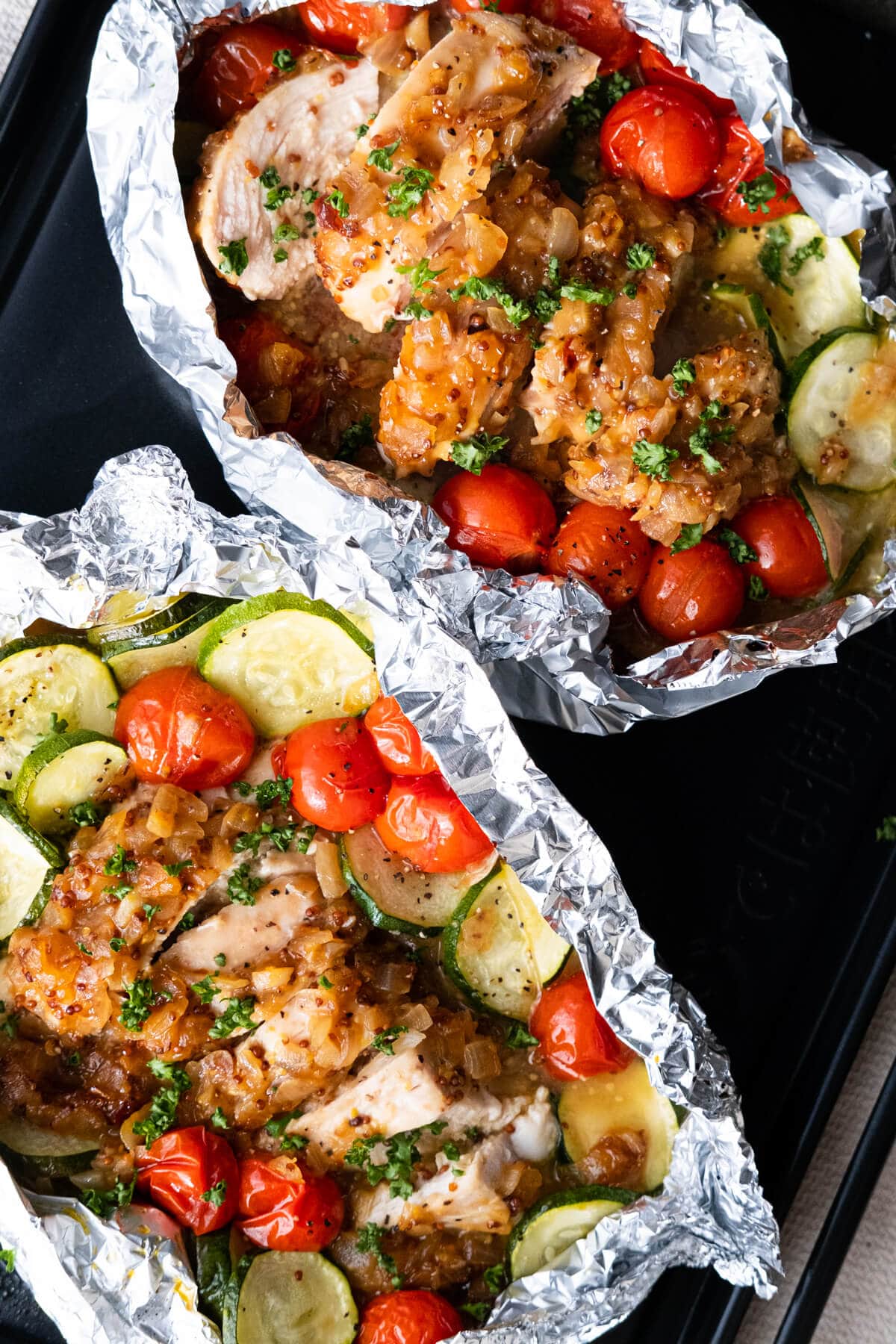 Two packs of baked chicken breasts in foil packets, topped with honey mustard sauce and cherry tomatoes, zucchini around. 