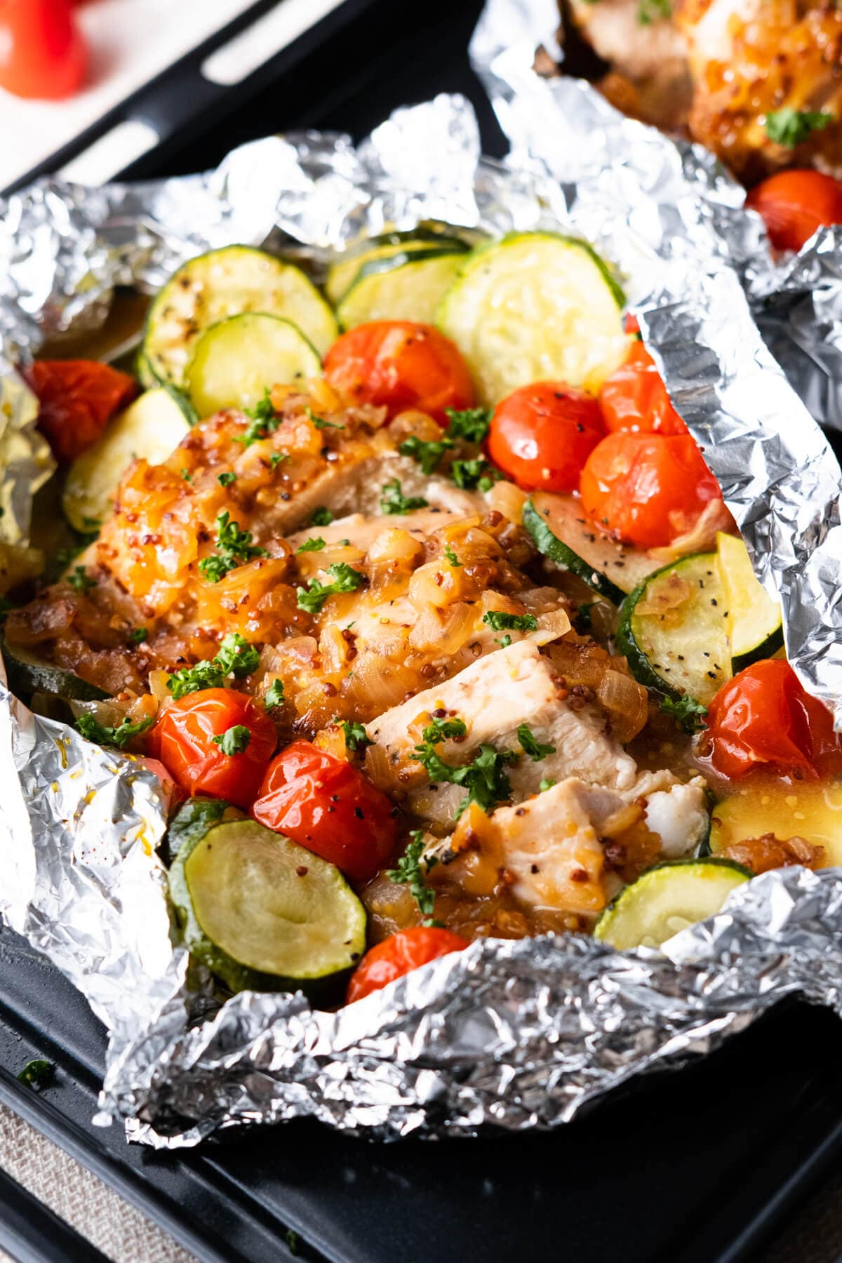 Close up on one foil packets filled with chicken breast, cherry tomatoes and zucchini, sprinkled with chopped parsley. 