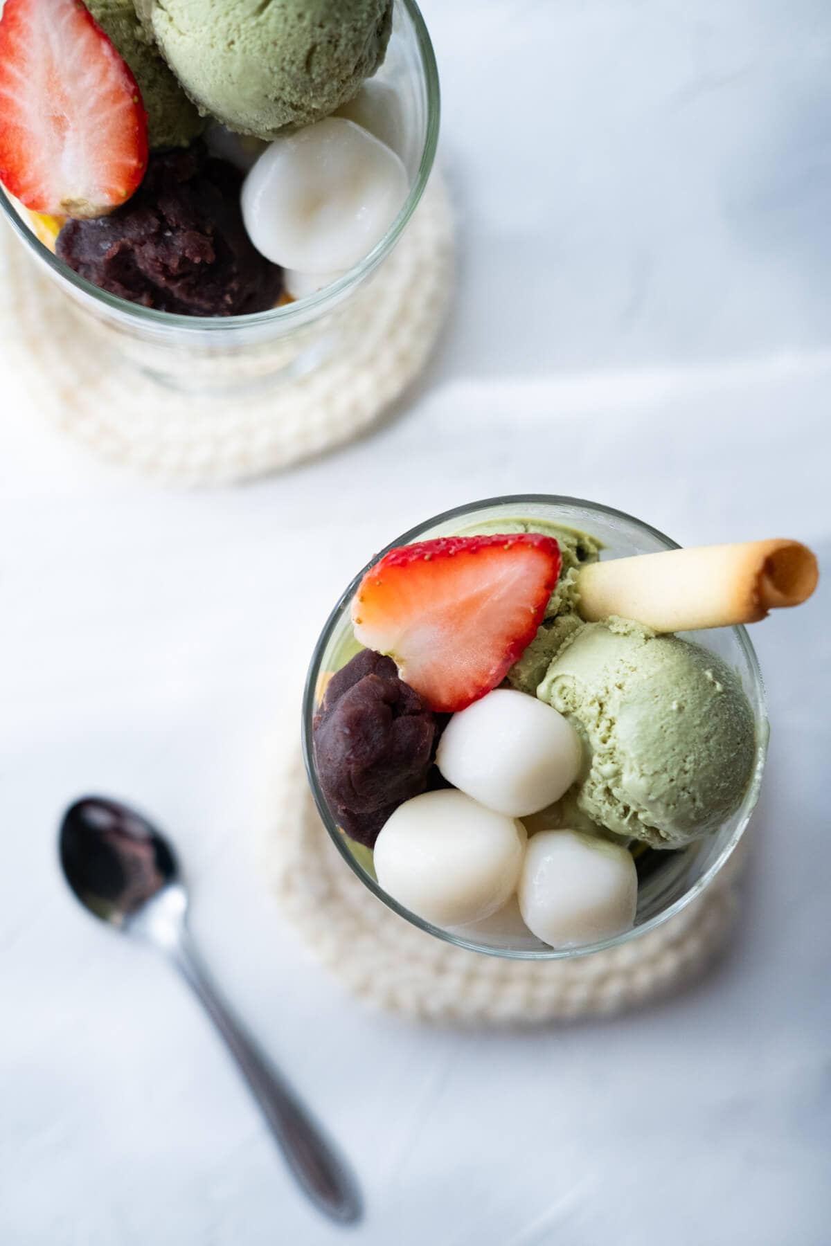 Two parfait glasses filled with matcha jelly, ice cream, mochi balls, sweet red bean paste, and strawberry halves. 