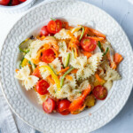 Pasta primavera is served in a white shallow plate and mixed with cherry tomato halves, farfalle, bell peppers, onion, carrots, and zucchini.