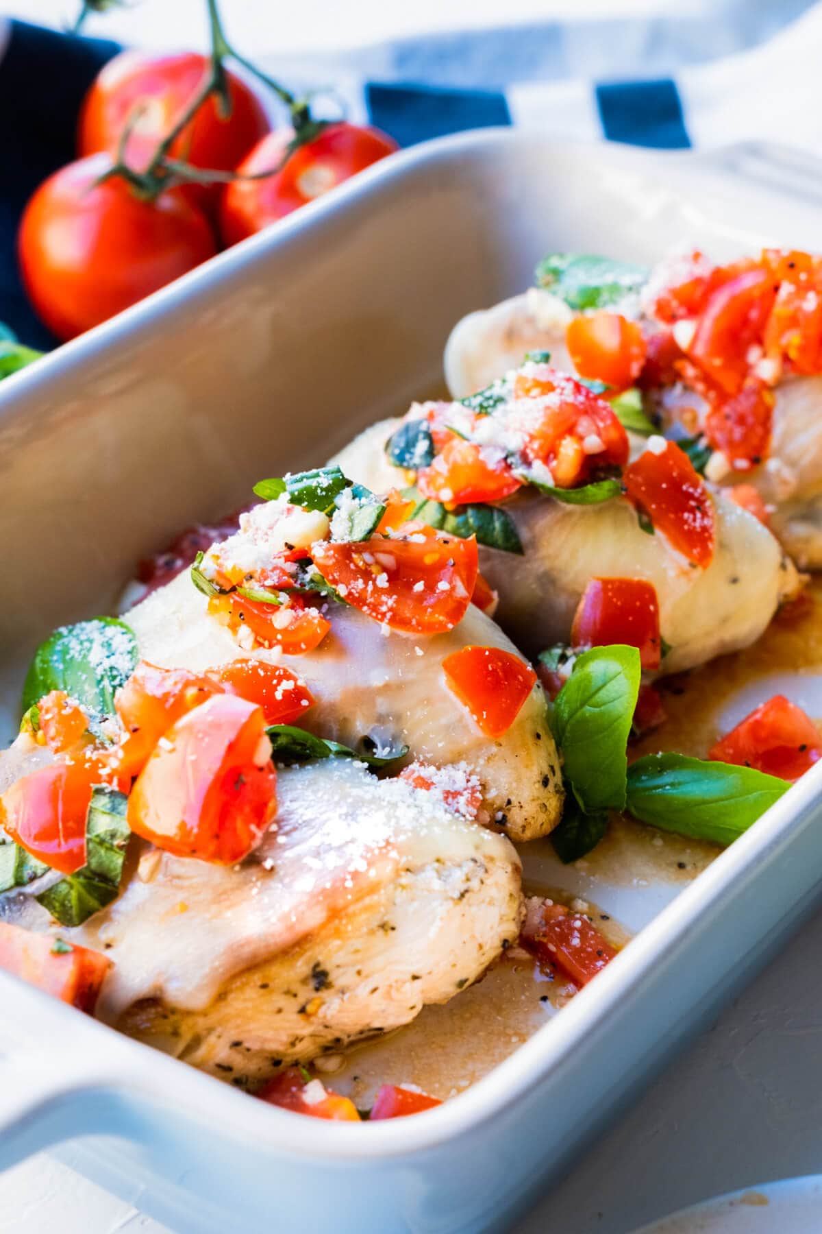 Chicken breast covered with melting cheese and served in a rectangular dish with chopped tomatoes and basil leaves.