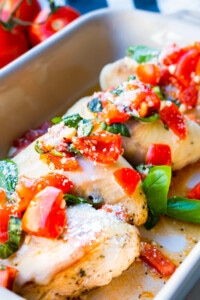 Chicken breast covered with melting cheese and served in a rectangular dish with chopped tomatoes and basil leaves.