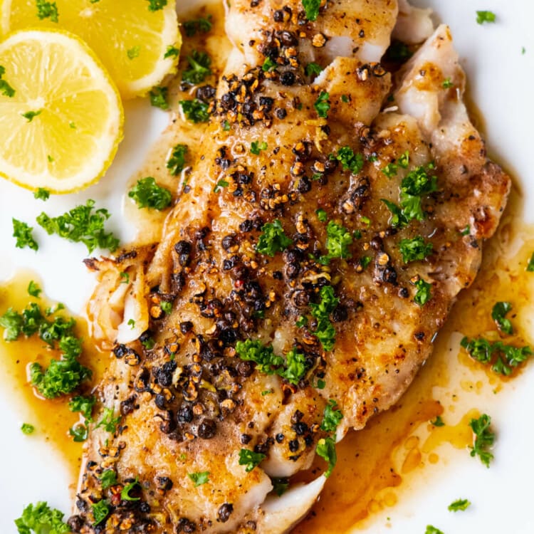 Pan-seared flounder fillet with buttery black pepper sauce served on a white shallow plate with lemon slices.