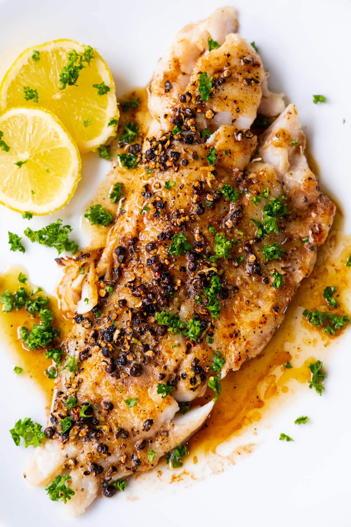 Pan-seared flounder fillet with buttery black pepper sauce and sprinkled with chopped parsley on top, alongside lemon slices. 