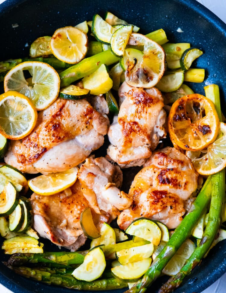 One pan greek chicken cooked in a skillet with asparagus, zucchini slices and lemon slices.