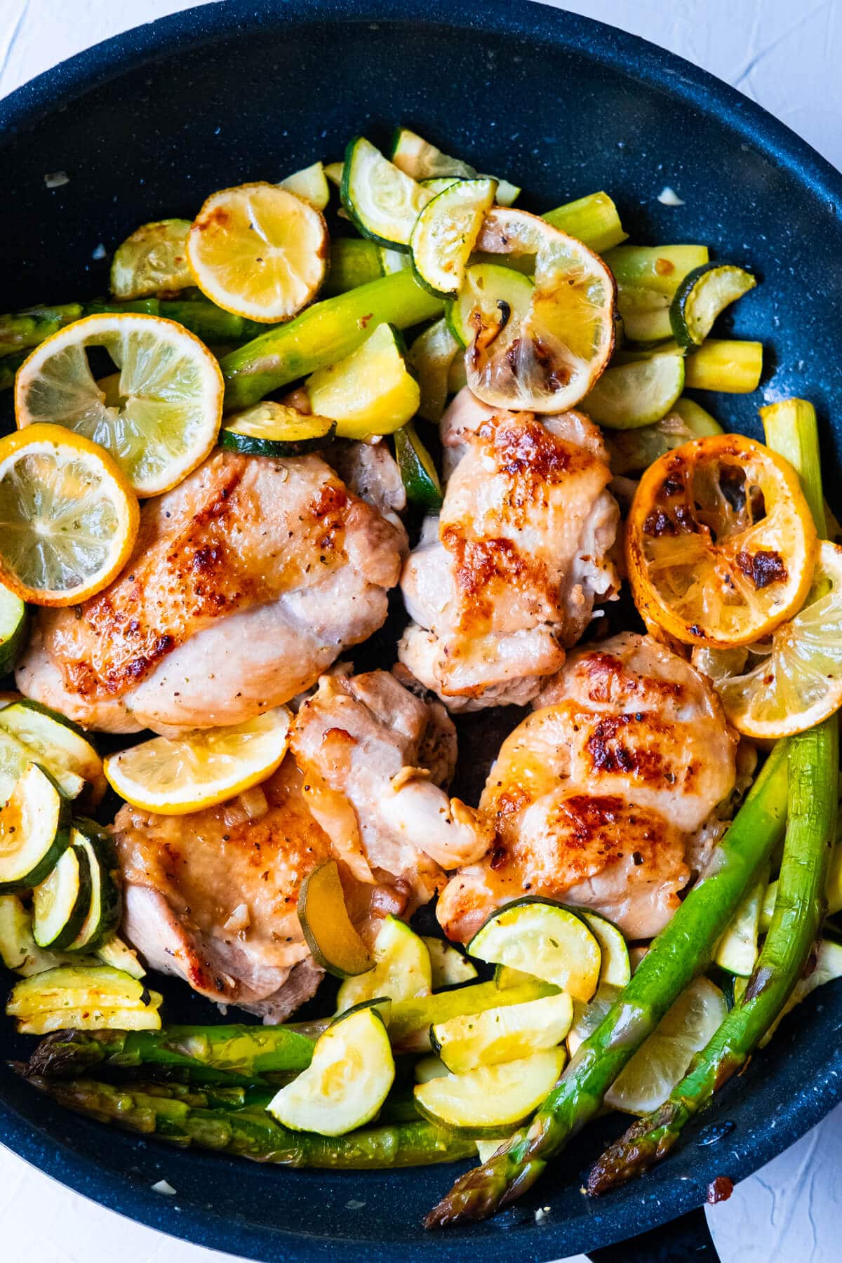 One pan greek chicken cooked in a skillet with asparagus, zucchini slices and lemon slices. 